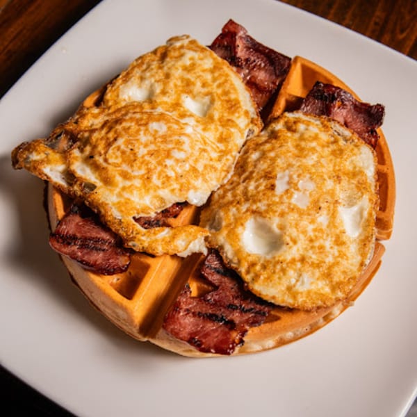 Two fried eggs and crispy bacon strips served on a golden waffle on a white plate.