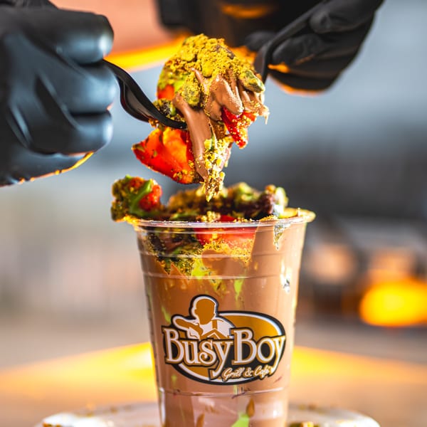 A hand in a black glove uses a fork to lift chocolate-covered strawberries and pistachios from a Busy Boy cup.