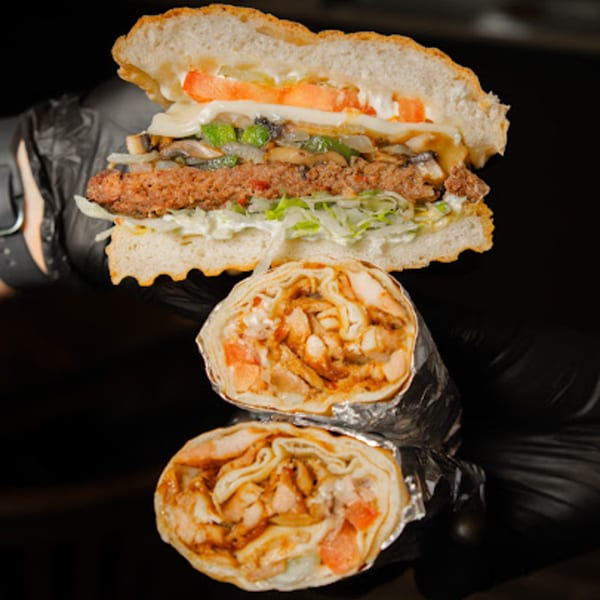 A large burger with vegetables and two chicken shawarma wraps held by a person in black gloves.
