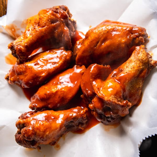 Six saucy, glistening chicken wings coated in a vibrant orange-red sauce, served on white parchment paper.