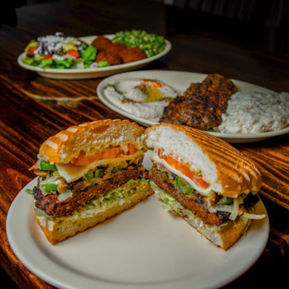 A cut Mediterranean sandwich on a white plate, with other dishes blurred in the background.