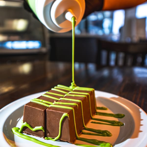 A hand drizzles green sauce over rectangular chocolate desserts on a white plate at Busy Boy Mediterranean Grill & Cafe.