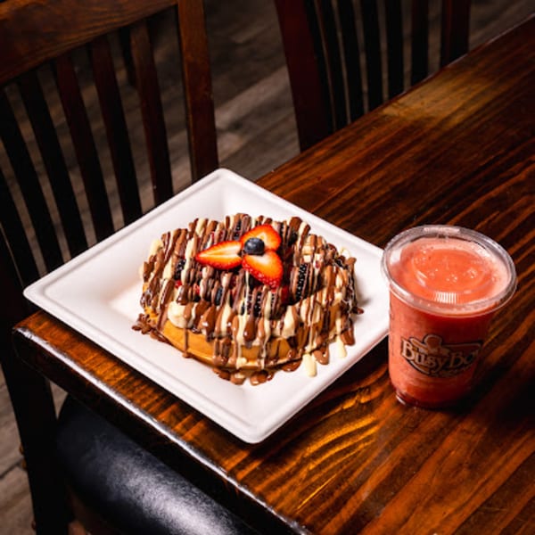 A waffle with chocolate and white chocolate drizzle, strawberries, and blueberries, next to a Busy Boy smoothie.