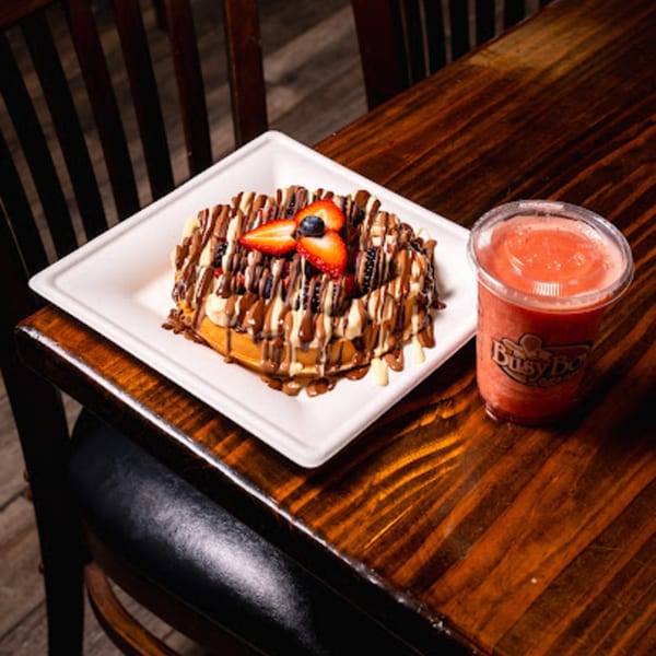 A waffle topped with chocolate, bananas, strawberries, and blueberries, next to a Busy Boy smoothie.