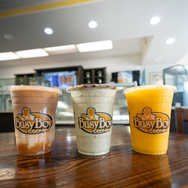 Three Busy Boy branded drinks, a caramel iced coffee, an Oreo shake, and an orange juice, on a wooden table.