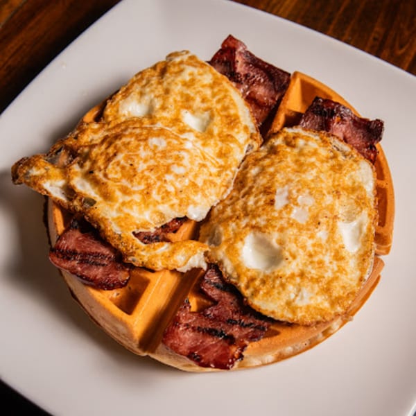 A waffle topped with two fried eggs and crispy bacon strips on a white plate.