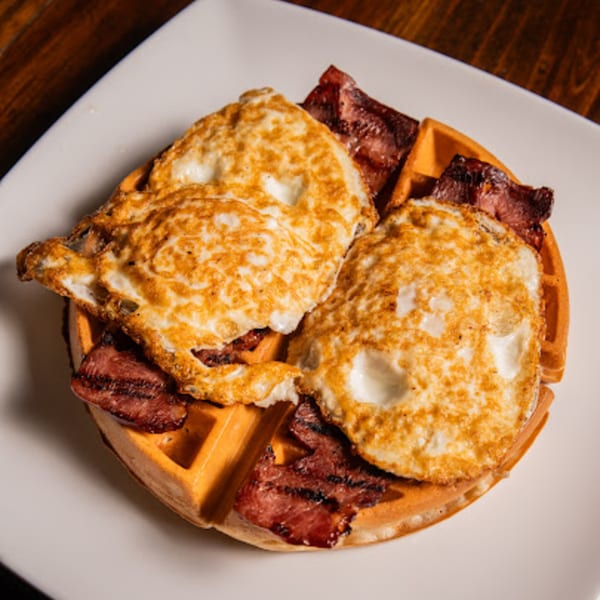 A golden waffle topped with crispy bacon and two fried eggs on a white plate.