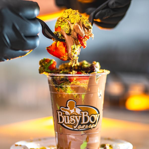 A hand in a black glove holds a forkful of chocolate, strawberries, and pistachios over a Busy Boy branded cup.